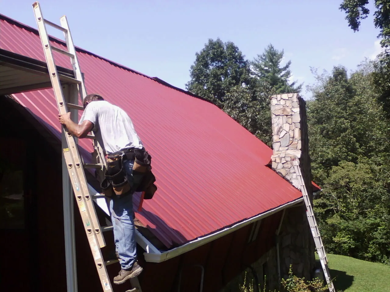 Roofer on a ladder beside a red metal roof and stone chimney — Soffit & Fascia Repair by Roofing Brothers in Sprague River